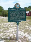 John Muir historical marker sign in a sandy field with trees and a small building