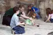 A group of friends including redheads in a sundress and blue jeans using digging tools in a sandy area.