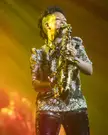English television presenter with short curly black hair playing a saxophone on stage in a floral print top