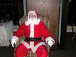A smiling, plus-size Santa Claus in a red suit with white fur trim sitting in a striped armchair.