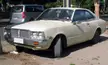 Toyota Corona vintage cream-colored coupe parked next to a silver minivan in a sunny outdoor area