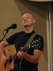 A bald man in a black t-shirt playing an acoustic guitar and singing into a microphone at Woodsong Coffee House.