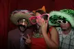 Three people in funny costumes with a sombrero, pink glasses, and a green fuzzy hat posing against a red curtain