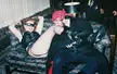 Paula in a black leather top and fishnets reclining on a floral sofa with a man in a red bandana