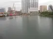 City skyline with tall buildings and construction cranes along a riverbank under an overcast sky.
