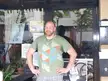 Muscular bald man in a green geometric t-shirt and plaid pants with a wrist tattoo standing by a storefront