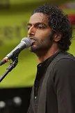 Chand, a man with curly dark hair and brown skin, singing into a microphone outdoors.