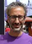 Baddiel wearing a purple t-shirt and black-rimmed glasses standing near a food stall