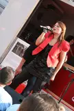 Annemie Coenen with red hair in a red jacket and black sequined mini dress singing into a microphone on a stage