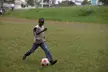 A person with dark skin wearing a tan t-shirt and blue denim jeans kicks a soccer ball on a grassy field.