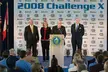 Four people in formal attire stand behind a podium at the 08 Challenge X event with US Department of Energy flags.