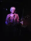 A blonde woman with pale skin in a red and blue plaid shirt playing an electric guitar on a dark stage.