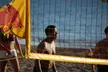 Three tanned people in a bikini and shorts standing by a volleyball net on a beach near the ocean.