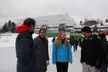 A group of friends, including blonde girls, smiling and talking on an outdoor ice rink in front of a white building.