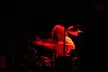Kanye West shirtless with curly hair playing a dw drum set under red lighting on a dark stage