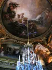 Palace of Versailles interior with a ceiling fresco, crystal chandelier, and people in classical drapery