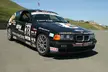 Black BMW race car with sponsor decals parked on an asphalt race track against a grassy hill.