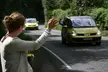 Rachel, with brown hair in a bun and a light cardigan, waves at two yellow cars on a tree-lined road.