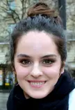 Noémie Merlant with dark brown hair in a high bun, wearing gold hoop earrings and a black scarf