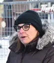 Filomena Tassi wearing a black beanie, glasses, and a brown winter coat with a faux-fur hood by a metal fence