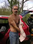 Yves, a shirtless man with a goatee and glasses, holding a white shirt by a red car