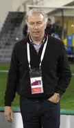 Gili as Umm Salal manager wearing a dark brown sweater and blue denim jeans on a soccer field