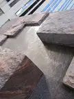 Close-up of water flowing over stacked reddish-brown stone slabs in front of a modern building