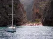 A white sailboat in a coastal cove in Mallorca, with a crowded beach and high rocky cliffs.