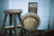 A wooden stool and bucket against a white tiled wall on a slatted floor in a liminal space.