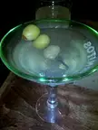 A martini glass with three green olives on a cocktail pick resting on a wooden table.