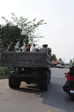 A group of people in casual clothing, including a white shirt, ride in the back of a grey truck followed by a motorcycle.