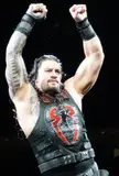 Roman Reigns with tribal tattoo sleeve and black tactical gear raising fists in a dark arena
