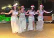 Four women in white sequined belly dancing costumes and red Santa hats posing outdoors at night with holiday lights