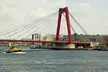 A red cable-stayed bridge over a river with a yellow tugboat and city skyline
