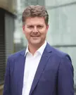 Andreas Schwab wearing a blue blazer and white shirt, smiling with brown wavy hair against a blurred building.