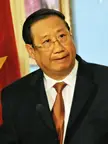 Phạm Gia Khiêm wearing glasses, a dark suit, and a brown tie in a professional portrait.