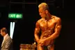 Brown, a muscular blonde bodybuilder with tanned skin posing on a competition stage next to a man in a grey suit