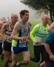 Athletic runners in singlets and short shorts competing in the West Yorkshire XC League on a foggy grassy field.