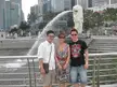 Three people in casual wear, including a white shirt and sundress, posing by the Merlion statue and city skyline.