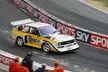 Stig Blomqvist driving a white and yellow Audi Quattro S1 rally car past a Sky Sports barrier at the Race of Champions.