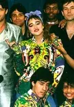 Madonna smiling in a colorful floral print shirt and purple headband, surrounded by dancers