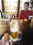 A smiling Chinese man in a red turtleneck sweater behind a bar counter with a glass of beer and a bun.