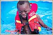 A smiling child with dark skin and short black hair wearing a red and yellow Body Glove life jacket in a swimming pool