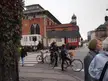 Group of people in leather jackets and denim jeans on bicycles crossing a city street by brick buildings